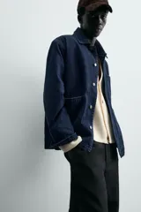 Chaqueta de denim de algodón en color azul oscuro con corte relaxed fit. Presenta cuello de solapa, cierre frontal con botones metálicos y tres bolsillos de parche en el delantero. Las mangas son largas con puños abotonados.