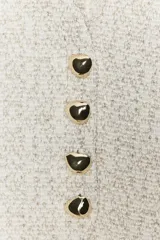 Chaleco de tejido texturado en tonos claros (blanco/beige/gris) con escote pico y cierre frontal de cuatro botones dorados. Presenta un bajo asimétrico.