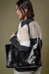 Bolso tipo tote de gran tamaño, confeccionado en cuero negro con paneles laterales de cuero charolado. Presenta costuras decorativas en hilo claro a lo largo de los laterales y doble asa de hombro.