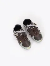 Zapatillas de cuero vacuno en color beige, con recortes de gamuza en la puntera y talón, doble cordón en tonos gris y marrón, hebilla lateral y dije colgante en la cordonera. Suela plana de goma.