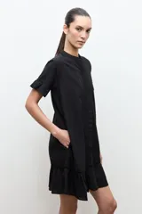 Vestido corto negro de corte amplio, con cuello mao, mangas cortas con volados y detalles de vainillas verticales en el frente. Cuenta con un volante fruncido en el ruedo.