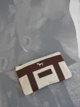 Sobre tipo cartuchera de lona naranja con detalles y apliques en cuero marrón. Presenta un aplique metálico en forma de caballo y un parche con el logo de la marca grabado en el frente.