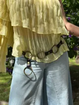 Cinturón negro de cuero con textura de cocodrilo, adornado con grandes aros metálicos dorados unidos por eslabones. Incluye una tira colgante con un aro grande en el extremo.