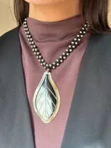 Collar con cordón de gamuza trenzado y cuentas de acero, con un gran dije central en forma de hoja hecho de guampa natural engarzado en alpaca.