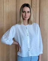 Camisa blanca de manga larga con puños elásticos, cuello redondo y cierre frontal con botones. Presenta un patrón floral texturizado.