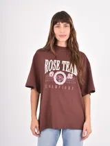 Remera de corte oversize color chocolate con estampa frontal estilo universitario en tonos rosados y blancos que incluye el texto Rose Team Champions.