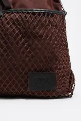 Bolso tipo tote de Bimba y Lola en color marrón, confeccionado en tejido técnico con un bolsillo frontal exterior cubierto por una red decorativa. Cuenta con asas superiores, correa ajustable extraíble y parche con logo de la marca en la parte inferior.