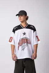 Camiseta blanca de poliéster con cuello en V negro y ribete rojo. Estampado frontal con una estrella negra y texto con efecto desgastado. Mangas cortas con números '0' y '1' en color rojo.