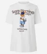 Remera blanca de algodón con cuello redondo y manga corta. Presenta un estampado central con texto que dice "Sunny Mornings Slow Days" y debajo un dibujo de un conejo con gorra azul, shorts y una mochila.