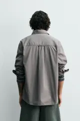 Camisa blanca de algodón con corte relajado, cuello solapa, manga larga con puño abotonado y bolsillo de plastrón en el pecho. Presenta bajo asimétrico con aberturas laterales y cierre frontal de botonadura.