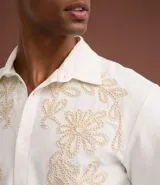 Camisa de corte comfort confeccionada en algodón, color blanco, con cuello solapa, manga corta y cierre frontal de botones. Presenta un delicado bordado floral en tono beige en la parte superior delantera.