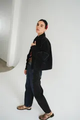 Campera de jean azul oscuro, estilo bomber, con cuello camisero y cierre frontal. Presenta un parche rectangular bordado en la espalda con patrón de rombos y texto.