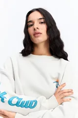 Buzo de algodón color marrón chocolate, con cuello redondo y diseño holgado. Presenta una estampa gráfica en la espalda con motivos de atardecer y montañas, junto con texto decorativo. Además, incluye una estampa en la manga con letras grandes.