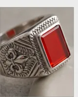 Anillo de acero tipo sello con piedra central rectangular de color rojo y diseño grabado en los laterales.
