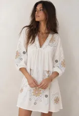 Vestido corto blanco de corte oversized, con escote en V y mangas 3/4 abullonadas. Presenta detalles bordados y estampados en tonos tierra y celeste en el frente y las mangas.
