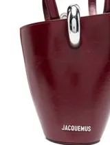 Bolso tipo cubo Jacquemus modelo Le Petit, confeccionado en cuero color bordó con acabado brillante. Cuenta con asas dobles superiores, correa bandolera extraíble, detalle de anilla metálica plateada en la parte frontal y el logotipo de la marca en letras metálicas.