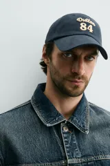 Gorra de seis paneles azul marino confeccionada en tejido de algodón, con bordado frontal en tono contrastante con la leyenda "button 84".