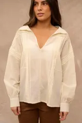 Blusa color crema de manga larga con detalles de alforzas verticales en el frente y hombros, escote en V con cuello mao y puños abotonados.