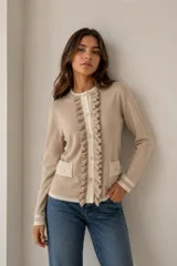Chaqueta de punto color beige con detalles en color crema, cuello redondo y cierre frontal con botones. Presenta volados decorativos a lo largo de la abertura frontal y dos bolsillos delanteros con solapa. Las mangas largas tienen puños con franjas horizontales.