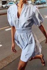 Vestido corto de color celeste con estampado floral, cuello redondo con volante y escote en pico. Presenta mangas cortas abullonadas con puños elásticos y cintura con nido de abeja elástico.