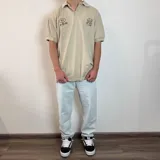 Chomba de corte oversize color beige, con cuello polo y cierre frontal de botones. Presenta bordados en el pecho con motivos de golf y tipografía cursiva.