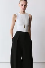 Pantalón negro de tiro alto, con pliegues en el delantero y bolsillos laterales ocultos. Cierre frontal con cremallera y ganchos metálicos.