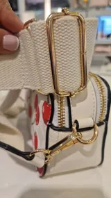 Cartera tipo bandolera de formato rectangular con estampado de cerezas sobre fondo claro. Cuenta con cierre superior, tirador con flecos y correa de tela ajustable y extraíble.