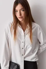 Camisa blanca de calce relajado, con cuello camisero, cierre frontal de botones, mangas largas y bolsillo aplicado en el pecho.