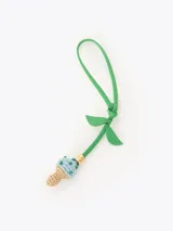 Charm para bolso con correa de cuero verde con forma de tallo y hojas, rematado con un dije en forma de hongo tejido a crochet en tonos beige y celeste con pequeñas cuentas decorativas y un detalle metálico dorado.