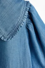 Blusa corta de denim liviano con mangas cortas abullonadas, cuello Peter Pan con volados y cierre frontal con tres tiras para atar.