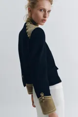 Chaqueta de estilo militar en color negro, con cuello subido y puños en terciopelo color verde oliva. Presenta detalles de alamares dorados en el frente y hombros, bolsillos con solapa y cierre frontal con botones metálicos en relieve.