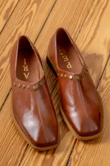 Zapato de cuero vacuno color marrón, con punta cuadrada y detalle de tachas metálicas en el borde superior. Presenta una costura central vertical y un taco geométrico.