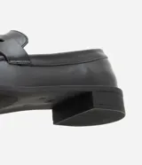 Mocasín Satinato de mujer confeccionado en cuero negro con acabado liso, puntera redondeada, costuras visibles, tira decorativa en la pala y suela con taco bajo.
