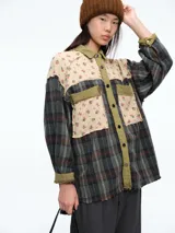 Camisa de corte oversize con diseño patchwork que combina paneles de tela a cuadros en tonos oscuros con paneles superiores de tela beige con estampado floral. Presenta cierre frontal con botones, cuello clásico y bolsillos delanteros con solapa.