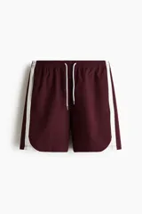 Short deportivo de nylon color bordó, con franjas laterales blancas, cintura elástica con cordón de ajuste blanco y bolsillos laterales.