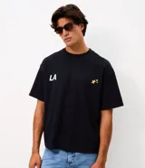 Remera de corte oversized confeccionada en mezcla de algodón y poliéster. Presenta cuello redondo, mangas cortas con hombros caídos y diseño estampado en la espalda con la frase 'LA THE START OF A NEW ERA' acompañada de estrellas amarillas.