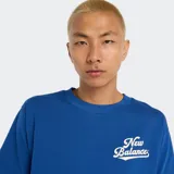 Remera de algodón azul vibrante, corte holgado, con cuello redondo y un pequeño estampado del logo de New Balance en tipografía cursiva blanca en el pecho.