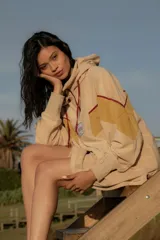 Hoodie de tela de toalla color beige, con capucha y cordones ajustables en color bordó. Presenta un diseño de bloques de color en tonos amarillos y bordó en la parte delantera y mangas, con un parche cuadrado con un logo en el pecho.