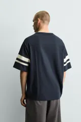 Camiseta de corte relajado, color azul marino, con cuello redondo y mangas cortas que presentan dos bandas horizontales en color crema. Cuenta con aberturas laterales en el bajo.