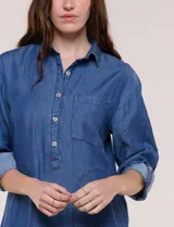 Vestido camisero blanco de rayón, con cuello con solapa, escote en V y pechera de abertura frontal con botones. Presenta un bolsillo delantero, bolsillos laterales aplicados y mangas largas con puño de largo regulable con presilla y botón. Cuenta con calce holgado y caída ligera, con ruedo irregular más largo en la parte trasera.
