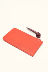 Monedero rectangular de cuero vacuno con textura granulada en color marrón topo. Cuenta con cierre superior metálico y un tirador decorativo de cuero en color naranja con un nudo. Presenta un pequeño logo grabado en el centro.