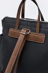 Mochila negra de poliéster texturizado con superficie acanalada. Cuenta con asas superiores marrones, tiras regulables para la espalda, bolsillo frontal con cierre metálico, compartimentos laterales y un interior amplio.