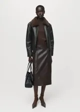Chaqueta efecto piel de color negro con diseño oversize, cuello camisero y forro interior de borreguito marrón. Cuenta con cierre frontal de cremallera, manga larga y dos bolsillos de parche laterales.