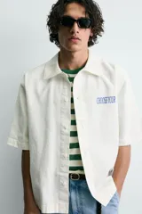 Camisa de corte relajado color verde oliva, con cuello solapa, manga corta y cierre frontal de botones. Presenta un estampado con el texto 'ASTRAL Drift' en el frente.