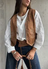 Chaleco de gamuza color camel con bordados decorativos en hilo claro y cierre frontal mediante lazos con borlas. Presenta un diseño de inspiración boho con terminaciones delicadas.