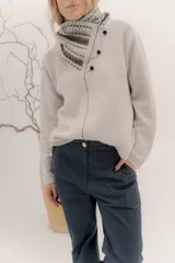 Polera tejida en lambswool color beige, con cuello alto y diseño de intarsia en tonos marrones y grises. Presenta botones forrados en el cuello para modular la apertura.