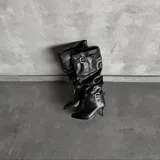 Botas altas negras de ecuero con diseño slouchy, tacón fino alto, puntera fina y tiras con hebillas metálicas ajustables en la caña y el tobillo.