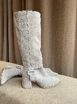 Botas de caña alta desmontables que se transforman en botinetas, confeccionadas en gamuza color beige. Presentan un diseño de punta semicuaadrada, taco cuadrado de 6 cm y detalle de estribo con hebilla forrada en el tobillo.