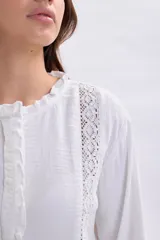 Blusa blanca de manga larga con detalles de encaje vertical en el frente, cuello con volados y terminación con volado en el ruedo. Presenta puños con volados y cierre frontal con botones.