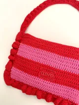 Cartera tipo shoulder bag tejida a crochet en hilo de algodón, con diseño a rayas en tonos de rojo y rosado, y detalles de volados en los bordes y la manija de hombro.